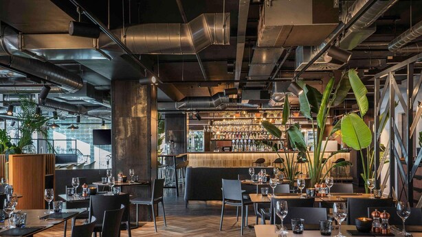 A modern restaurant with exposed ductwork, wooden flooring, and a bar area with a variety of bottles. - Mercure Newport Hotel (Newport)