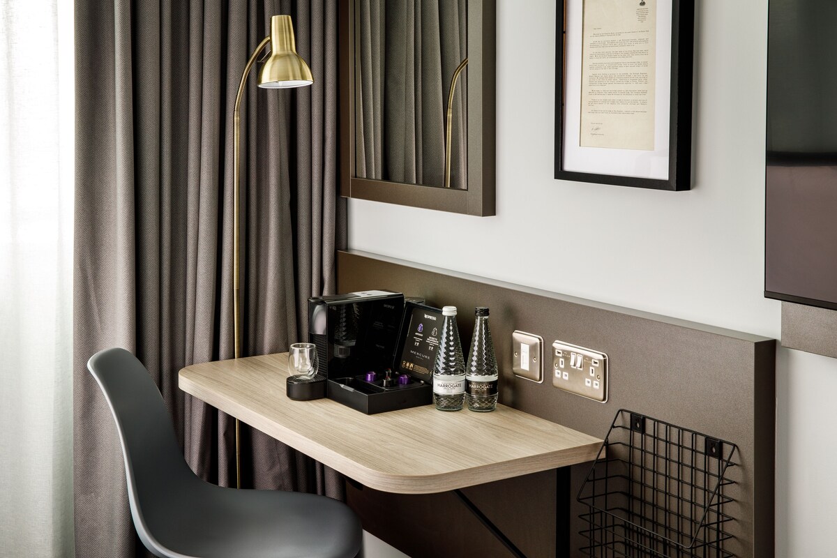 A modern hotel room with a wooden desk, a black chair, a framed menu, and a wall-mounted lamp.