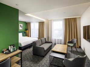 Ein Hotelzimmer mit Bett, Sofa, zwei Sessel, einem kleinen Tisch und einem Schreibtisch mit Frühstücksgebäck.