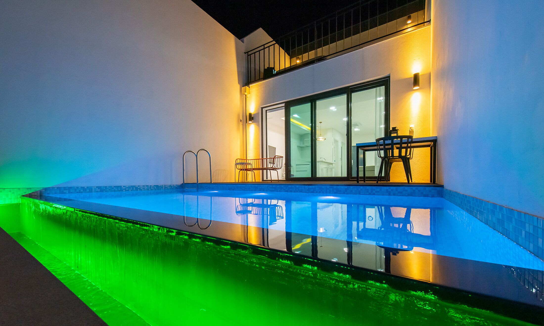 A modern outdoor swimming pool with a glass bottom, adjacent to a building with a dining area and a staircase.
