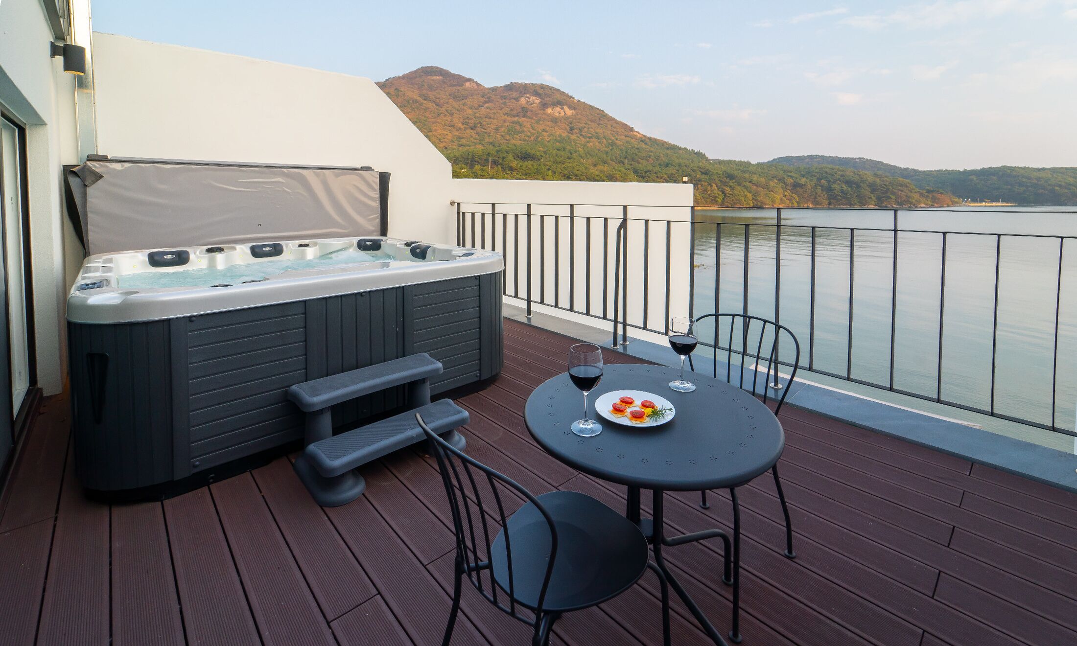 A balcony with a hot tub, a table with chairs, and a view of a lake and mountains.