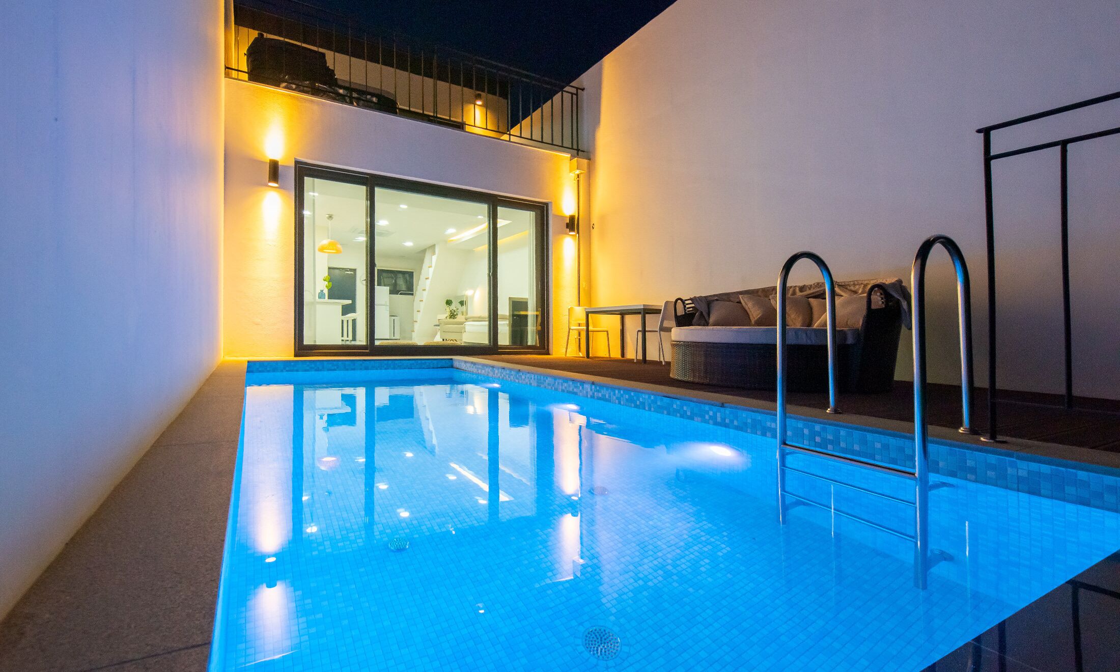 A swimming pool with a glass wall, a sofa, and a table on the patio.