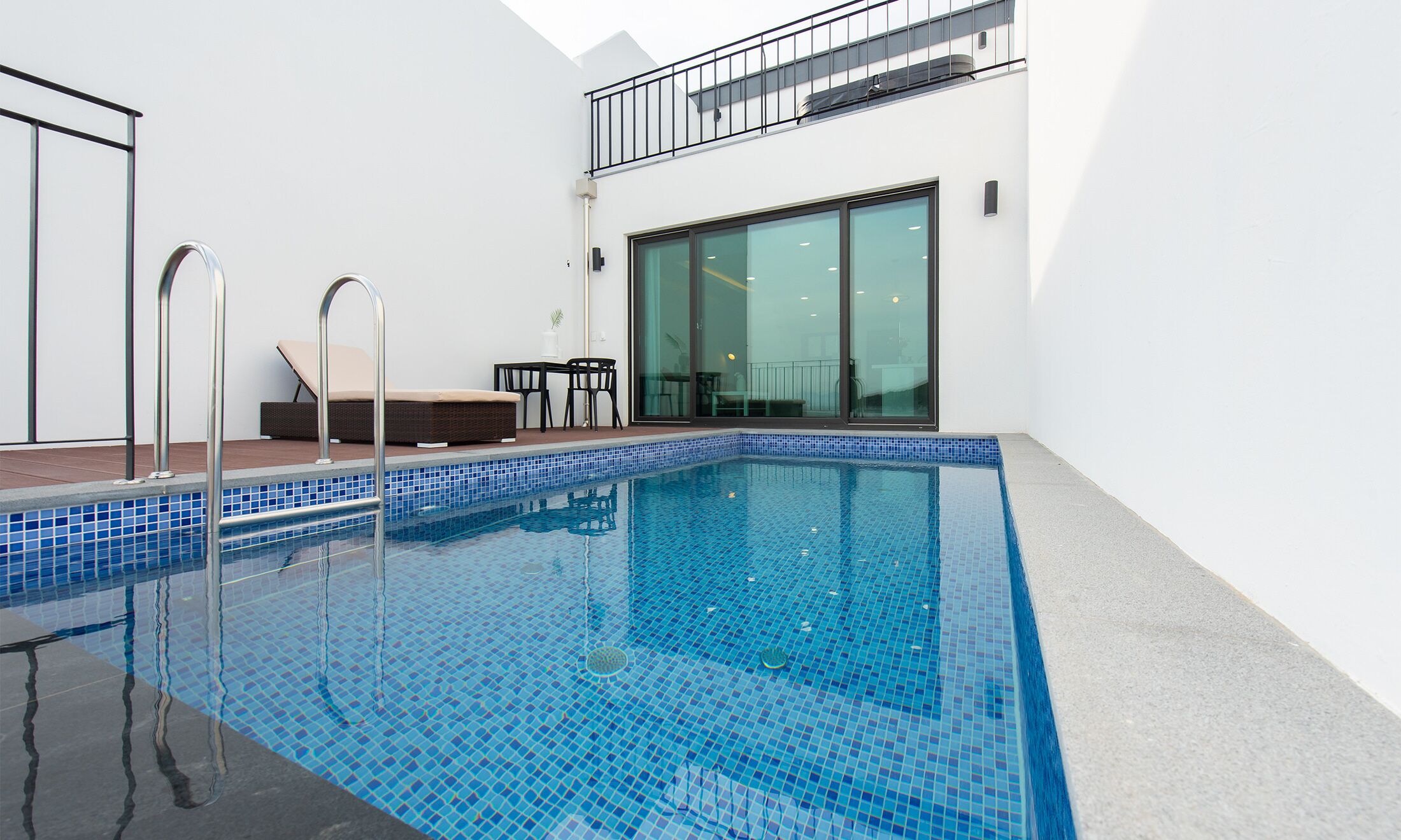 A modern indoor swimming pool with clear blue tiles, a sun lounger, and a seating area with a table and chairs.
