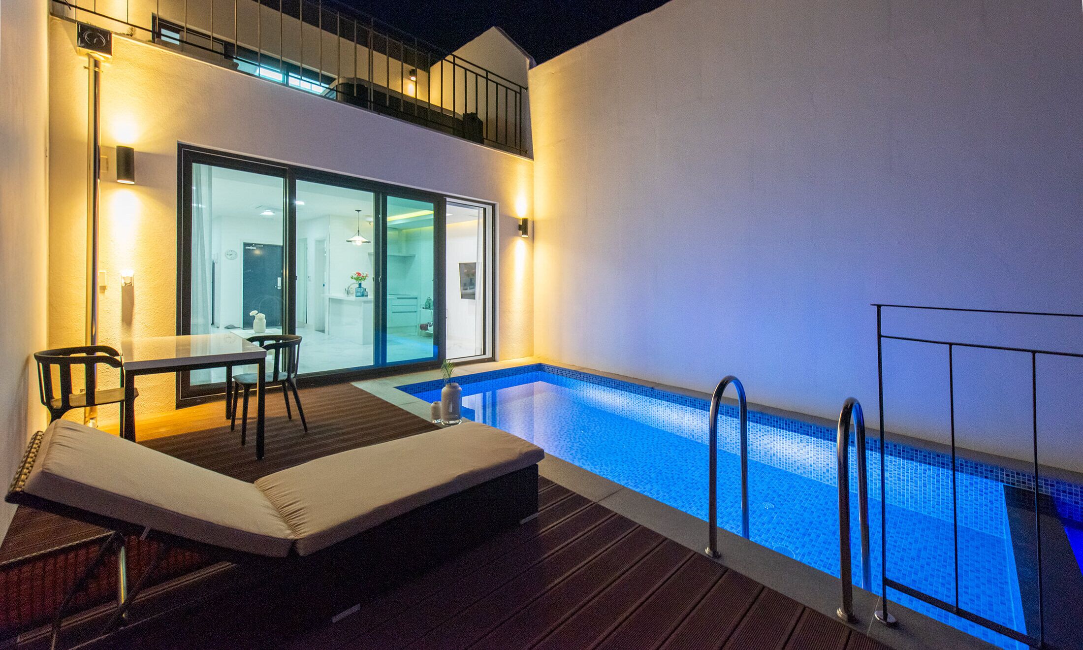 A modern outdoor pool area with a glass-enclosed room, a dining table, and a lounge chair.