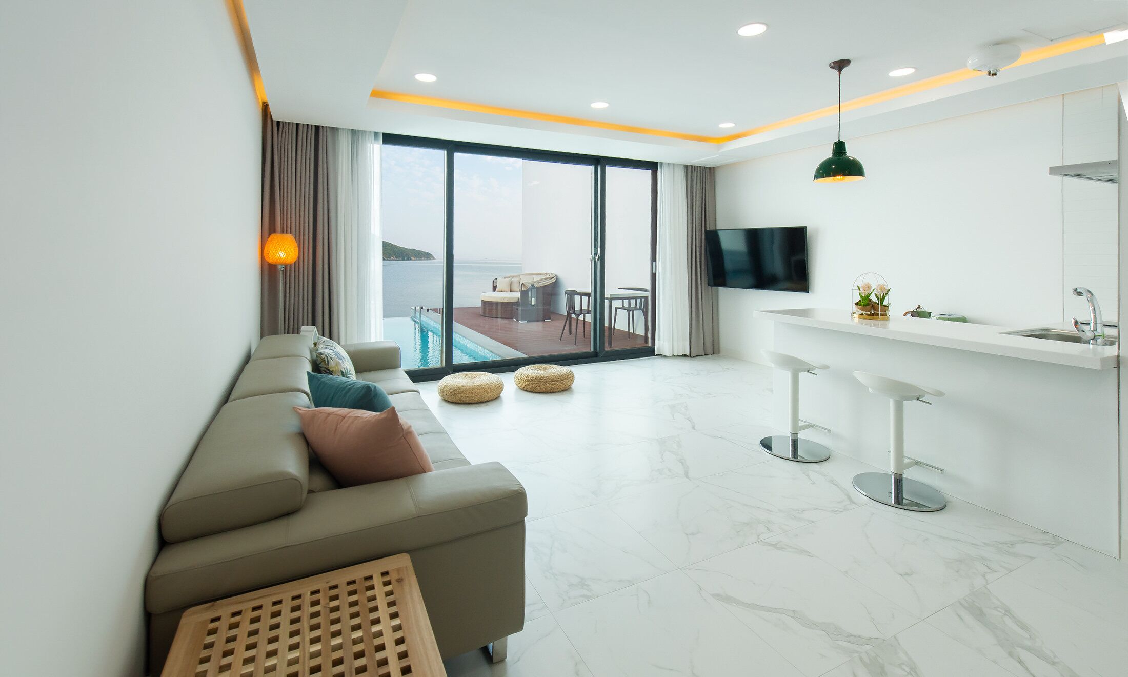 A modern living room with a sofa, a TV, a kitchen area, and a balcony with a pool view.