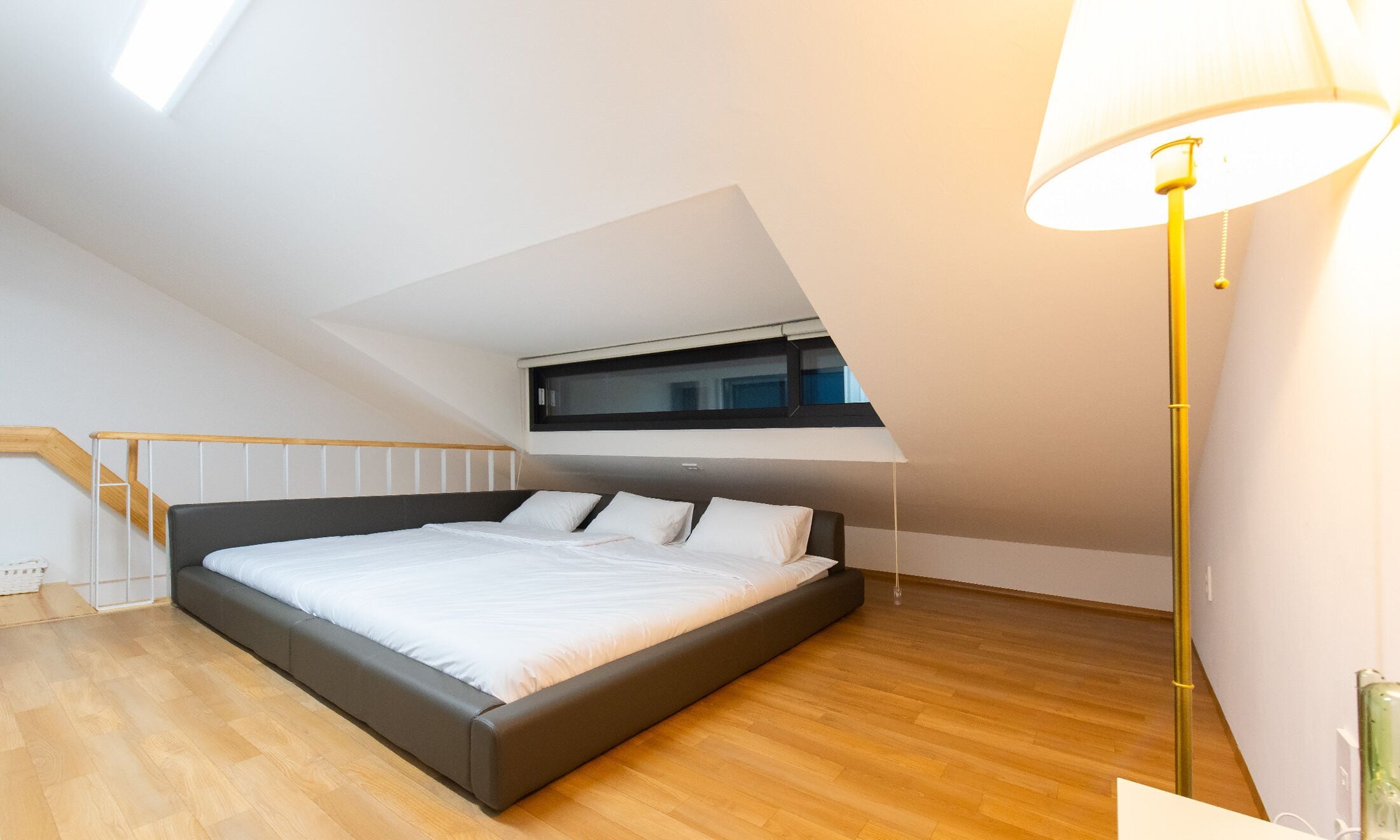 A modern bedroom with a large bed, wooden flooring, a skylight, and a floor lamp.