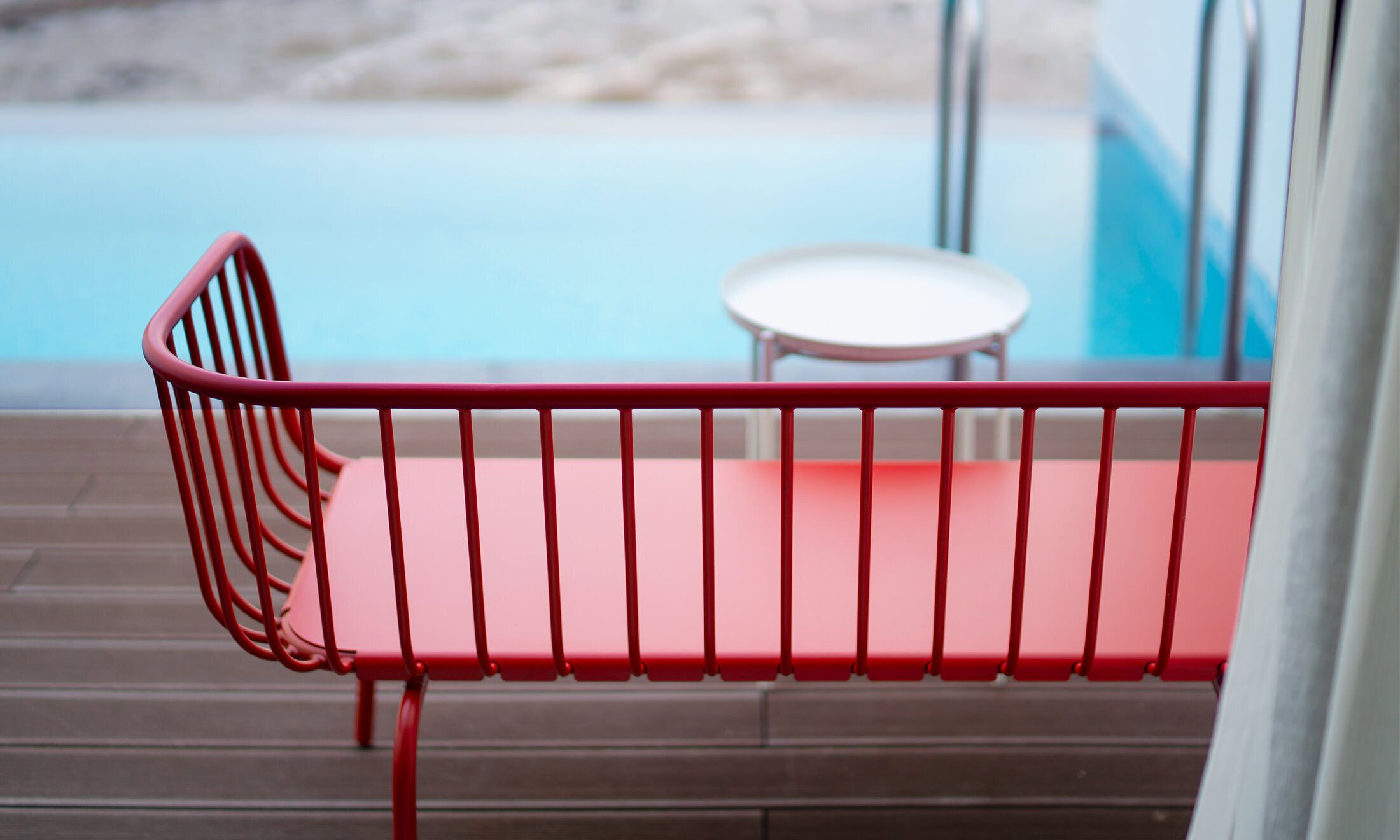 A red metal bench with a white cushion, positioned on a wooden deck overlooking a pool.