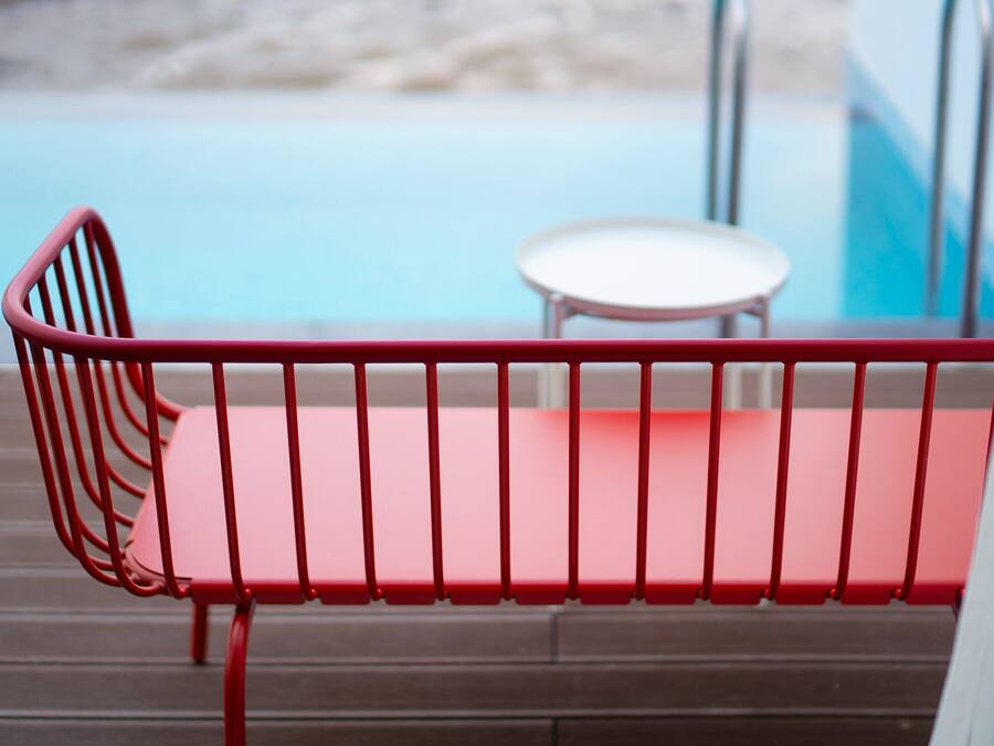 A red metal bench with a white cushion, positioned on a wooden deck overlooking a pool.