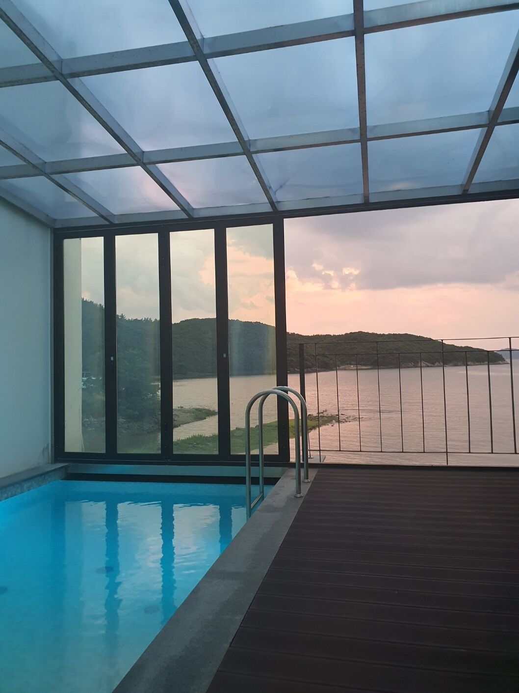 Ein Hallenbad mit Glasveranda, einer Metallleiter und Blick auf einen See und Hügel.