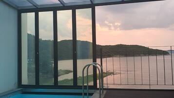 Ein Hallenbad mit Glasveranda, einer Metallleiter und Blick auf einen See und Hügel.