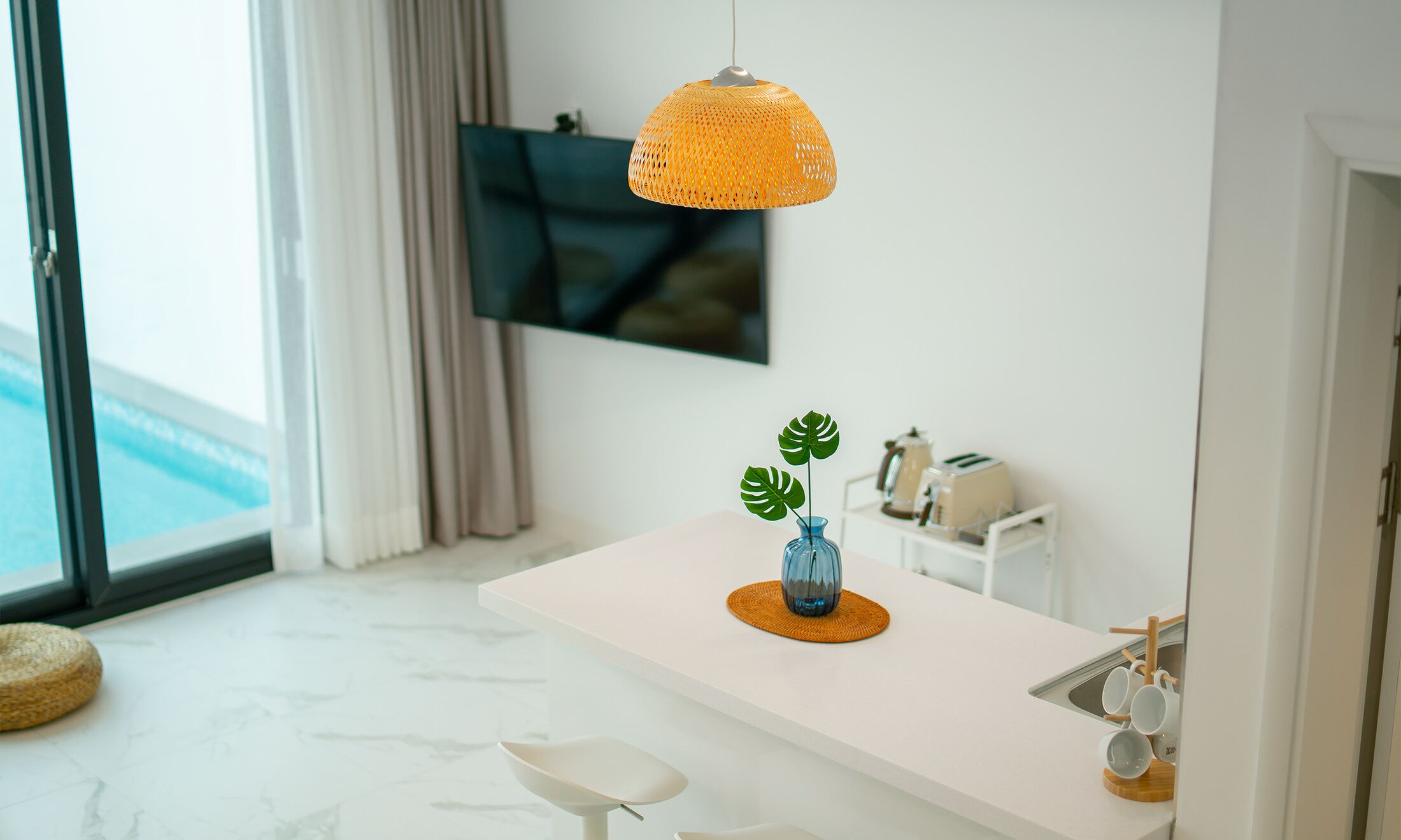 A modern kitchen with a white countertop, a hanging orange pendant light, a small blue vase with a plant, and a TV mounted on the wall.