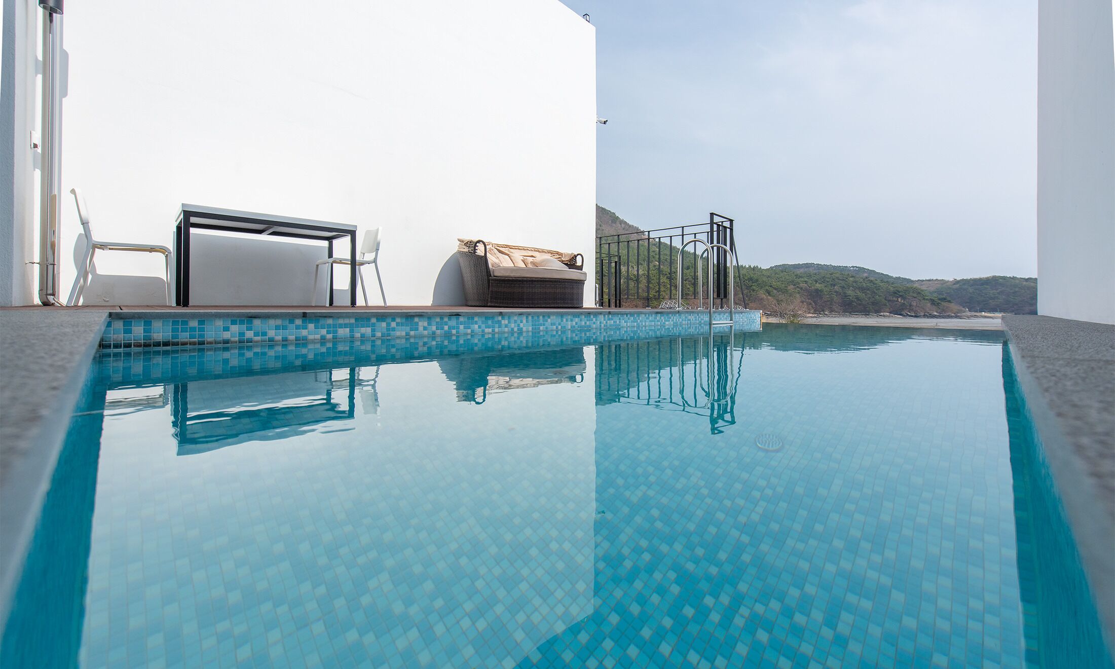 Ein klarer, blauer Infinity-Pool mit einer gefliesten Kante, ein Korbsessel, ein weißer Stuhl und eine kleine, überdachte Sitzgelegenheit.