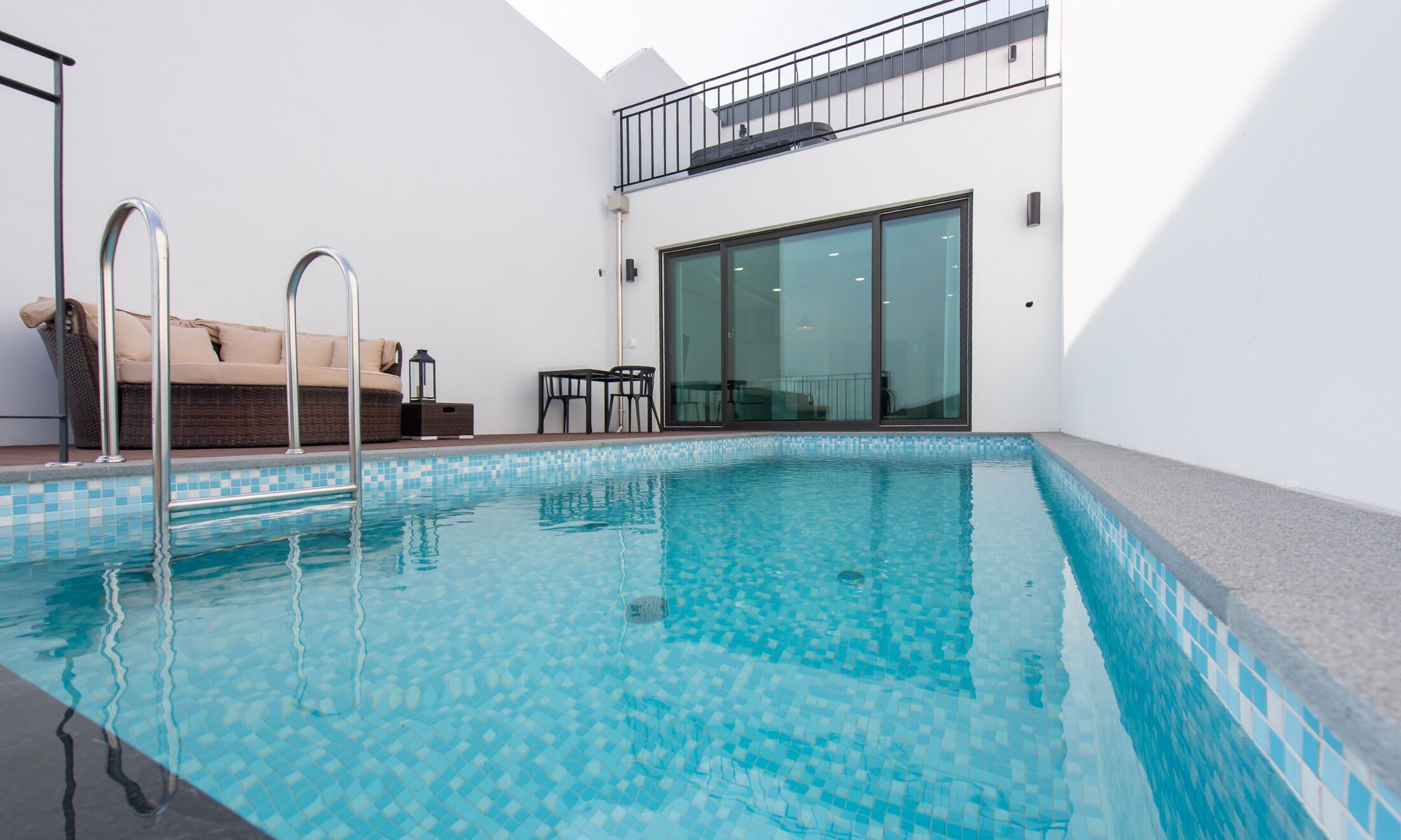 A modern indoor swimming pool with clear blue water, a metal ladder, a wicker sofa, and a glass door leading to a balcony.