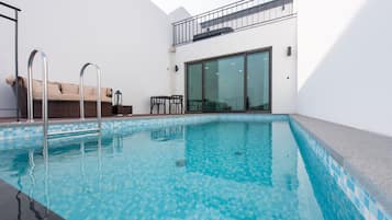 A modern indoor swimming pool with clear blue water, a metal ladder, a wicker sofa, and a glass door leading to a balcony.