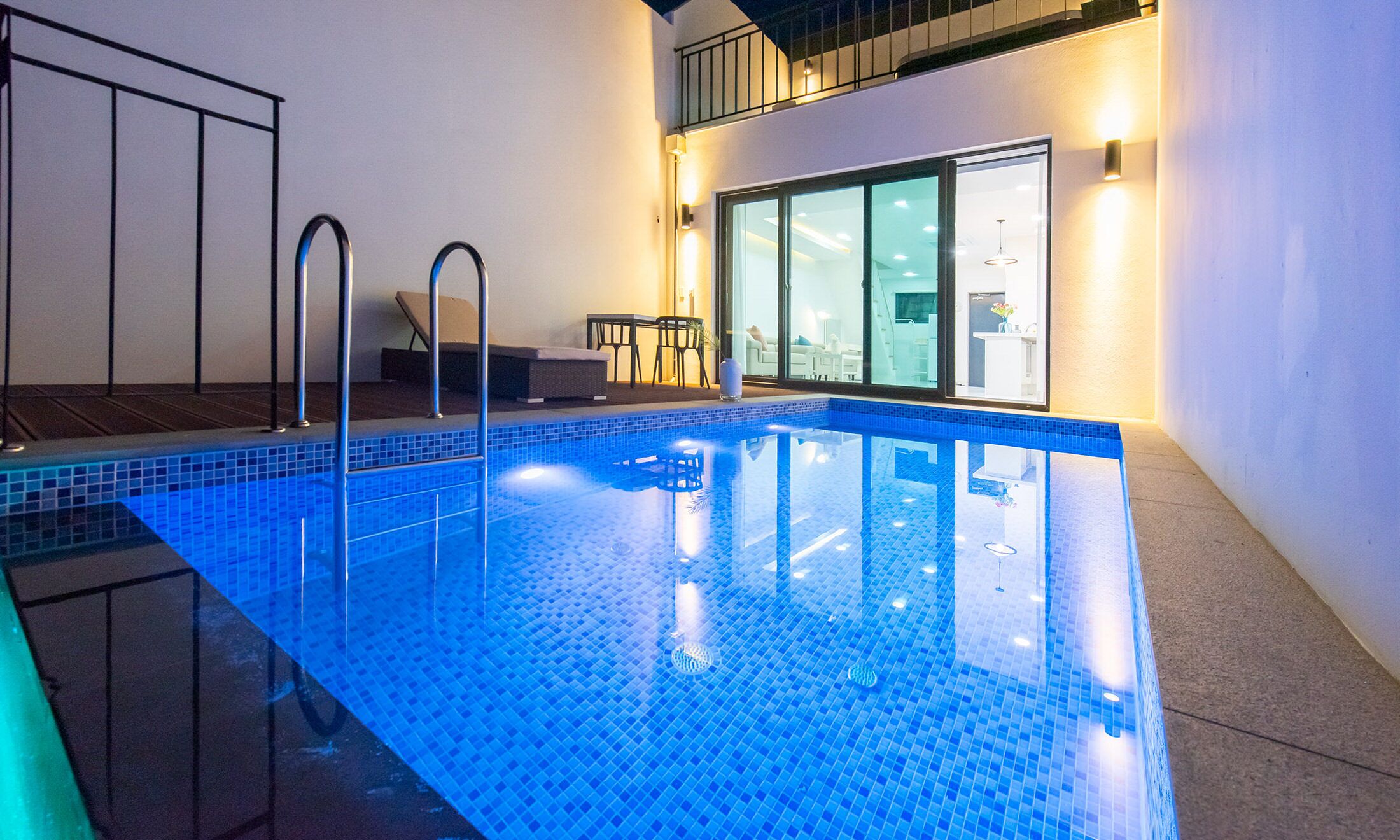 Ein moderner, überdachter Pool mit klaren, blauen Fliesen, eine Sonnenterrasse mit Sitzgelegenheiten und eine Glastür zu einem hellen Raum.
