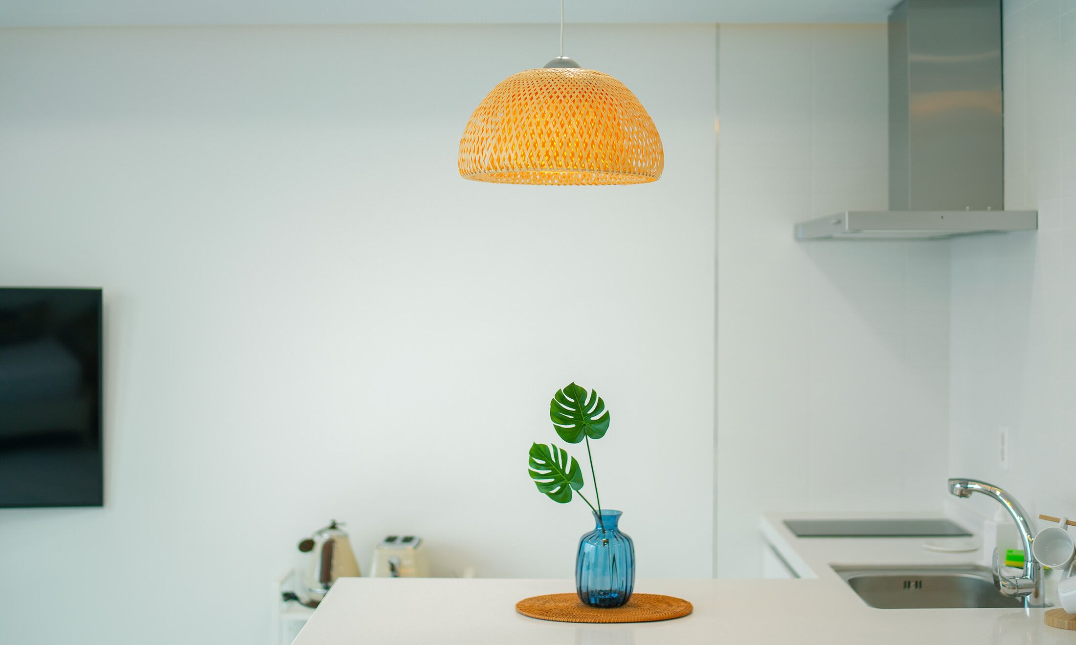 A modern kitchen with a hanging orange woven light fixture, a blue vase with a plant, and a stainless steel sink.