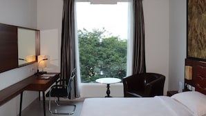 Quarto de hotel com cama, escrivaninha, luminária, cadeira, mesinha e janela com cortinas.