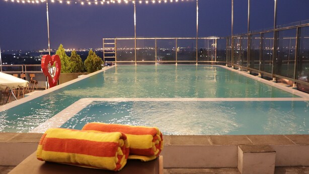 Uma piscina no terraço com água azul cristalina, rodeada por grades de vidro e vista para a cidade ao fundo.