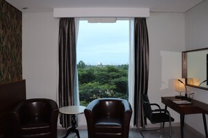 A hotel room with a large window, two leather chairs, a small round table, a desk with a lamp, and a mirror.