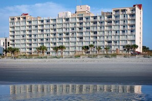 Un hôtel à plusieurs étages doté de balcons, situé près d’une plage bordée de palmiers.