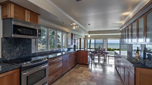 A modern kitchen with granite countertops, stainless steel appliances, and wooden cabinets. It features large windows offering a view of the ocean.