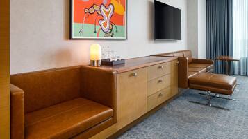 A hotel room with a brown leather sofa, a wooden side table with a lamp, a flat-screen TV, and a large window with curtains.