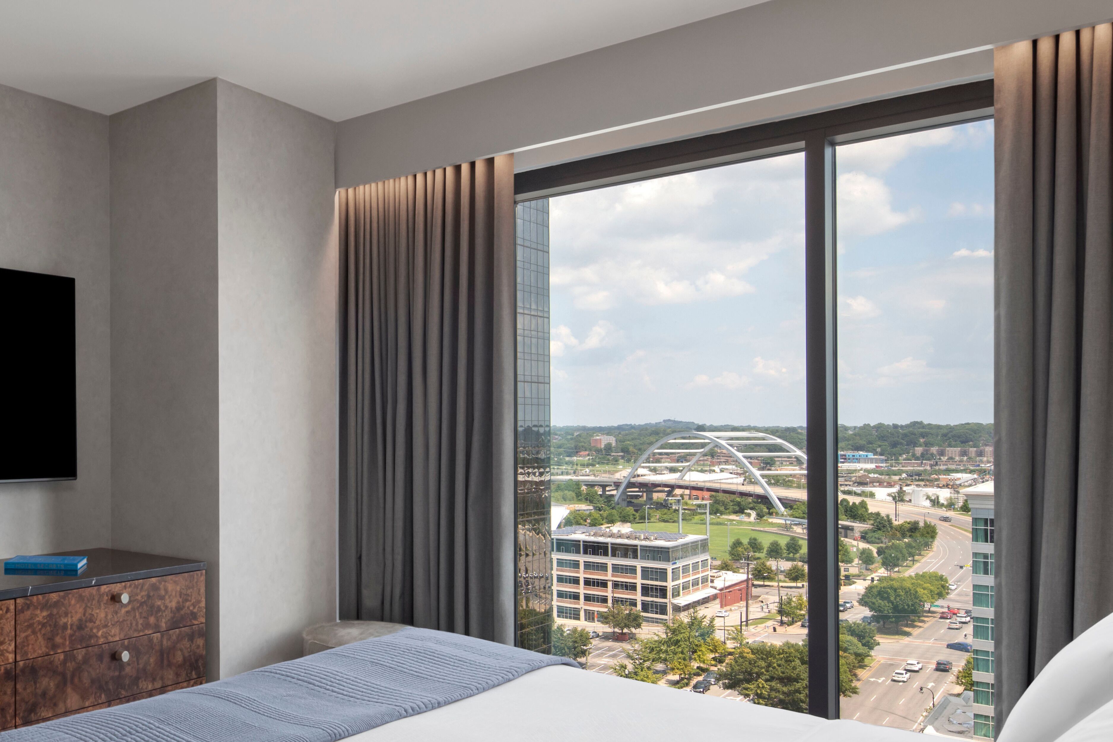 A hotel room with a large window, a bed, a flat-screen TV, and a wooden dresser.