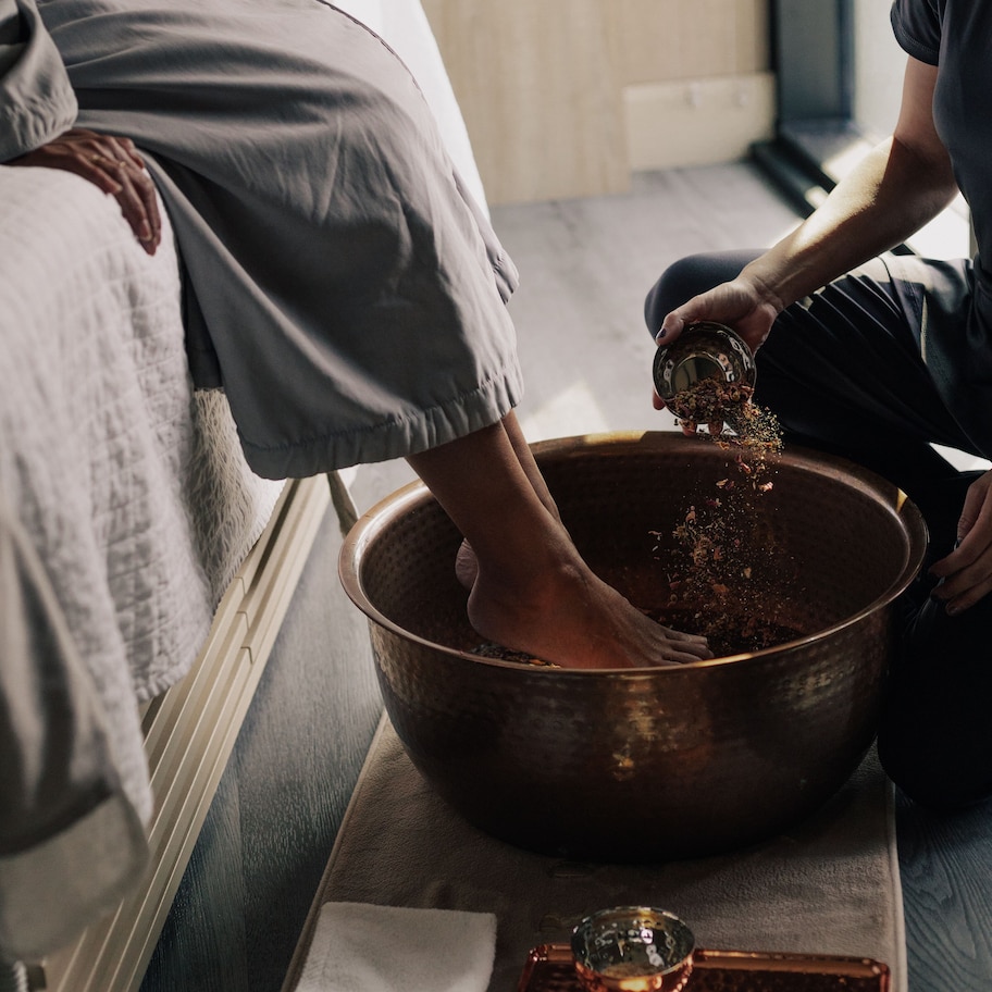 A person sitting on a bed, pouring a liquid from a glass into a copper bowl on a tray.