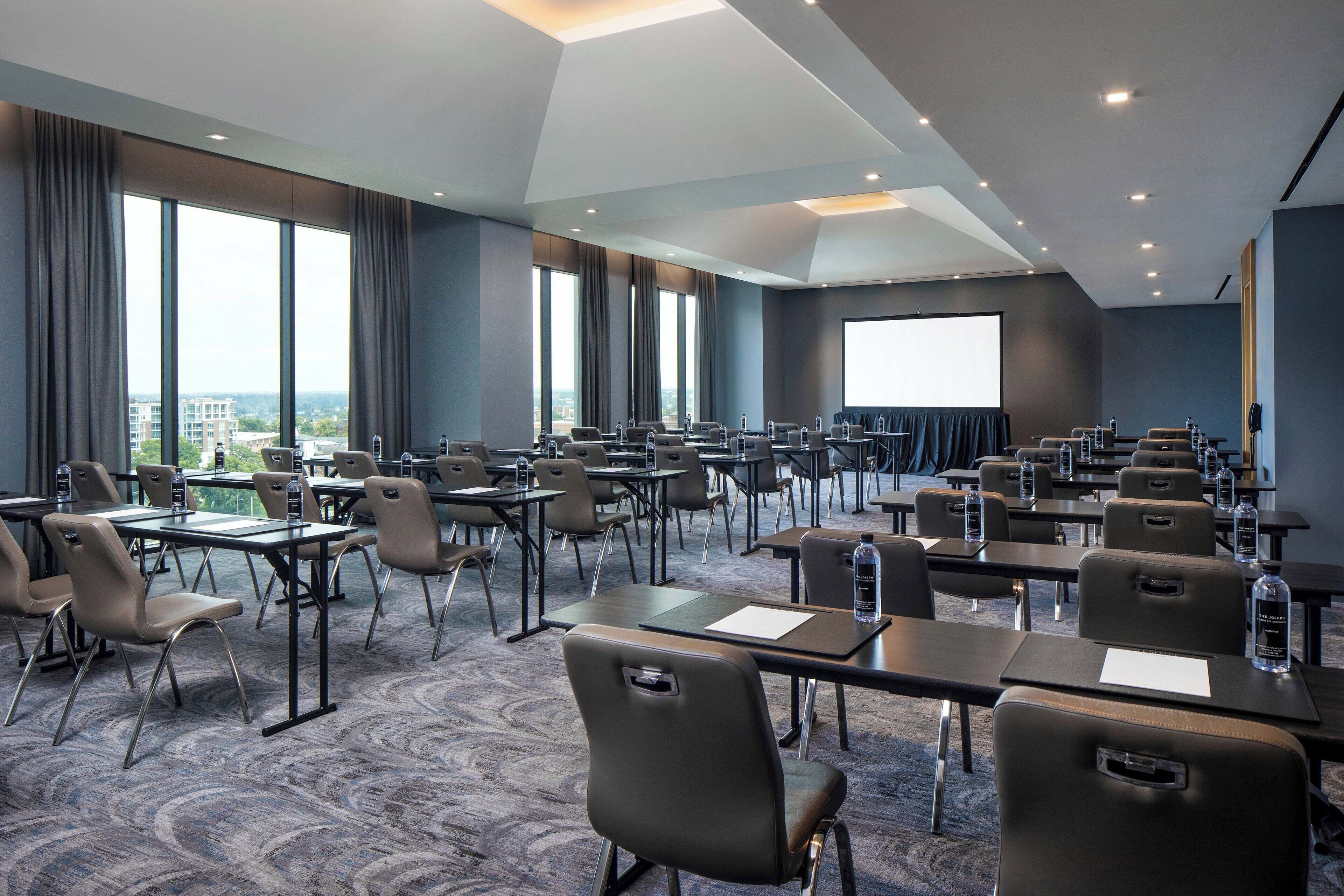 A conference room with multiple tables and chairs, large windows, and a projection screen.
