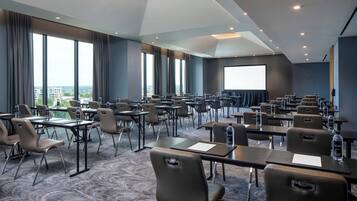 A conference room with multiple tables and chairs, large windows, and a projection screen.