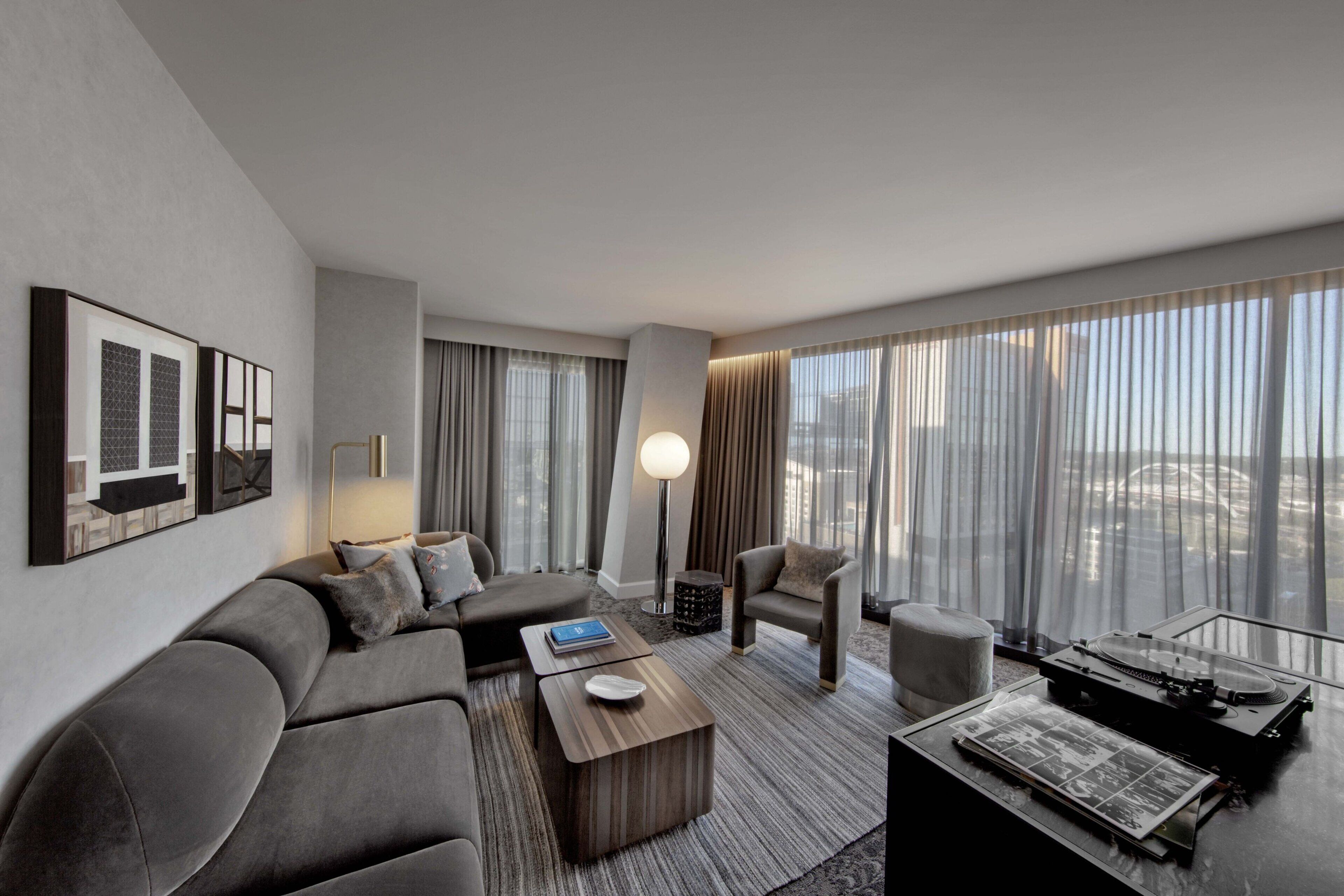 A modern hotel room with a grey sofa, a small armchair, a coffee table, and a large window with curtains.