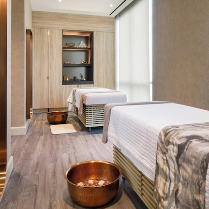 A modern spa room with two beds, a wooden floor, a large window, and a shelf with decorative items.