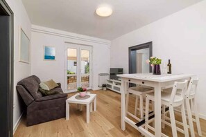 A compact living space with a sofa, a small table with a plant, a dining area with chairs, a TV, and a large window with a view.