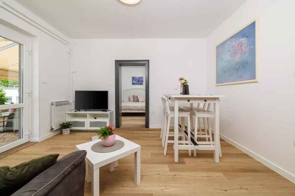 Un salon moderne doté d’un téléviseur à écran plat, d’un coin repas avec des chaises blanches, d’un canapé et d’une petite table sur laquelle est posée une plante en pot.