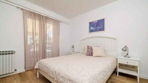 Quarto com cama, mesa de cabeceira, luminária, radiador e janela com cortinas.