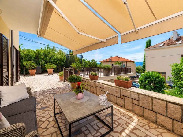 A covered outdoor area with a stone floor, a table, and chairs, surrounded by potted plants and a view of a house and trees.
