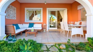 Un'accogliente area salotto esterna con divano, sedie e tavolo, immersa in un giardino e incorniciata da un arco bianco.