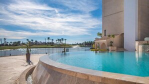 A swimming pool with a curved edge, a large building with a flat roof, and a clear sky with some clouds.