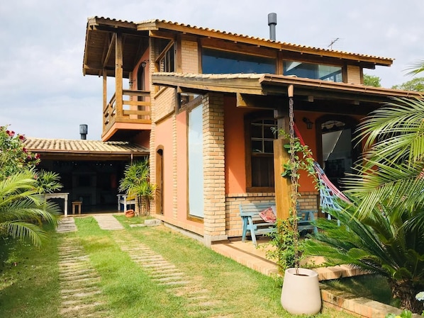 Uma casa de dois andares com varanda, alpendre coberto e um pequeno jardim com palmeiras e uma rede.