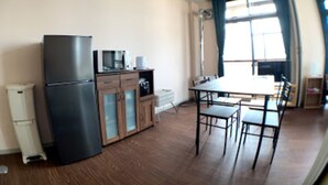 A compact kitchen with a refrigerator, microwave, and cabinets. There is a dining table with chairs and a window with curtains. - Big Stone Tsukuda 201 (Aomori)