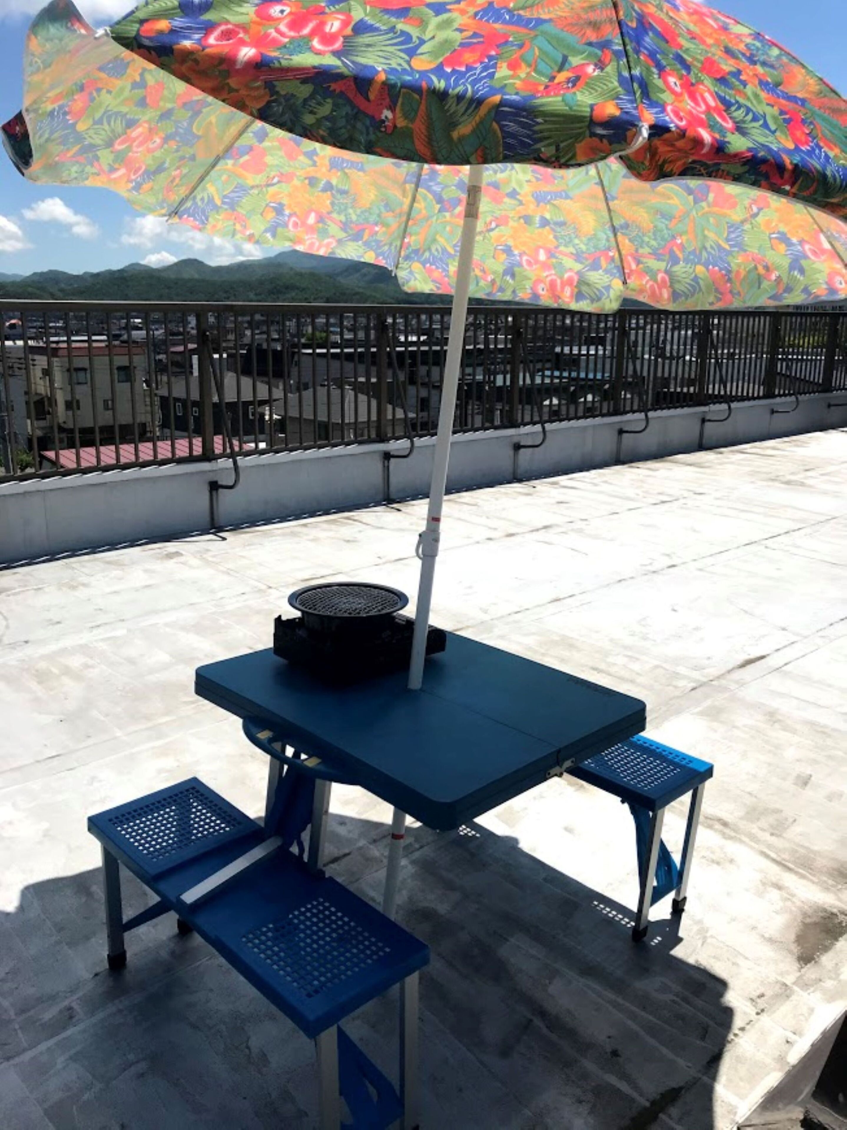 Une table bleue pour l’extérieur, surmontée d’un parasol coloré, deux bancs bleus et un barbecue posé sur la table.