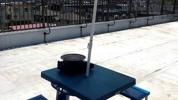 Une table bleue pour l’extérieur, surmontée d’un parasol coloré, deux bancs bleus et un barbecue posé sur la table.