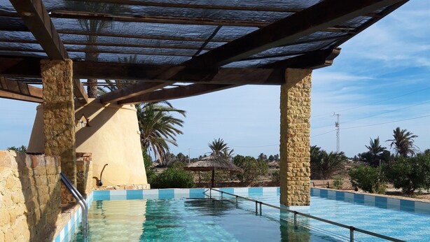 A clear swimming pool with stone walls and a wooden pergola overhead. - Bella Djerba (Sidi Salem)