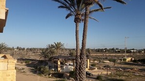 A building with a stone facade, a palm tree in the foreground, and a dry landscape with sparse vegetation and scattered structures in the background. - Bella Djerba (Sidi Salem)