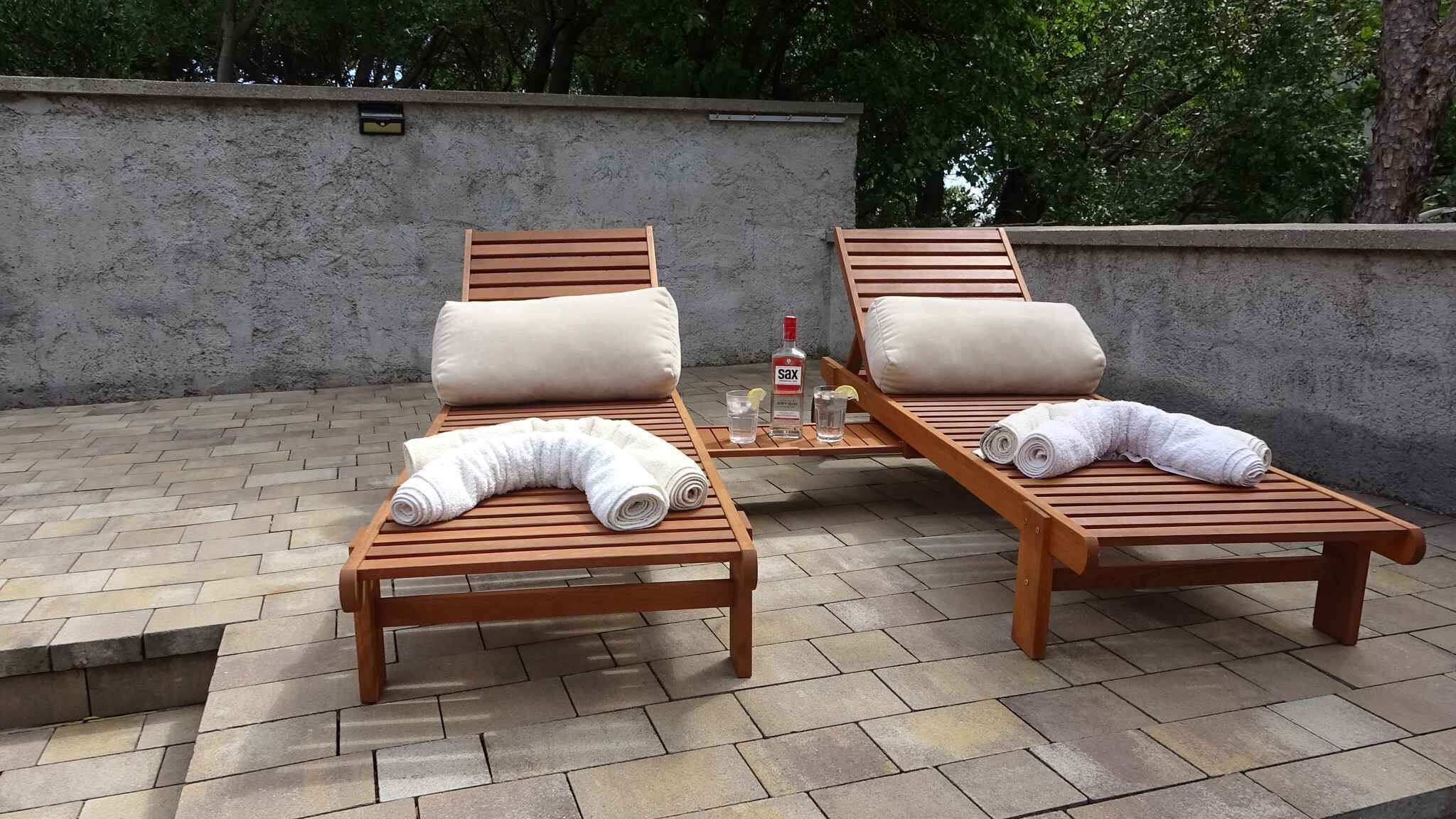 Two wooden lounge chairs with beige cushions, a bottle of Sax alcohol, and glasses on a small table.