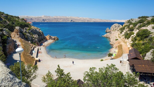 Une plage côtière aux eaux bleues et limpides, avec un terrain rocheux et quelques bâtiments, dont une petite construction surmontée d’un toit rouge.