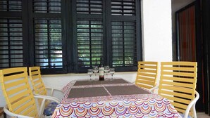 Une table dressée avec des verres à vin et une bouteille, entourée de chaises jaunes, avec une nappe à motifs et de grandes fenêtres en arrière-plan.
