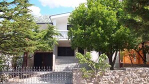 Une maison à deux étages, dotée d’un mur en pierre et d’un portail en métal, entourée d’arbres et baignée par un ciel dégagé.