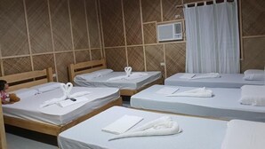 Una habitación con cuatro camas de madera, cada una con ropa blanca de cama y toallas. Hay una ventana con cortinas blancas y un equipo de aire acondicionado en la pared.