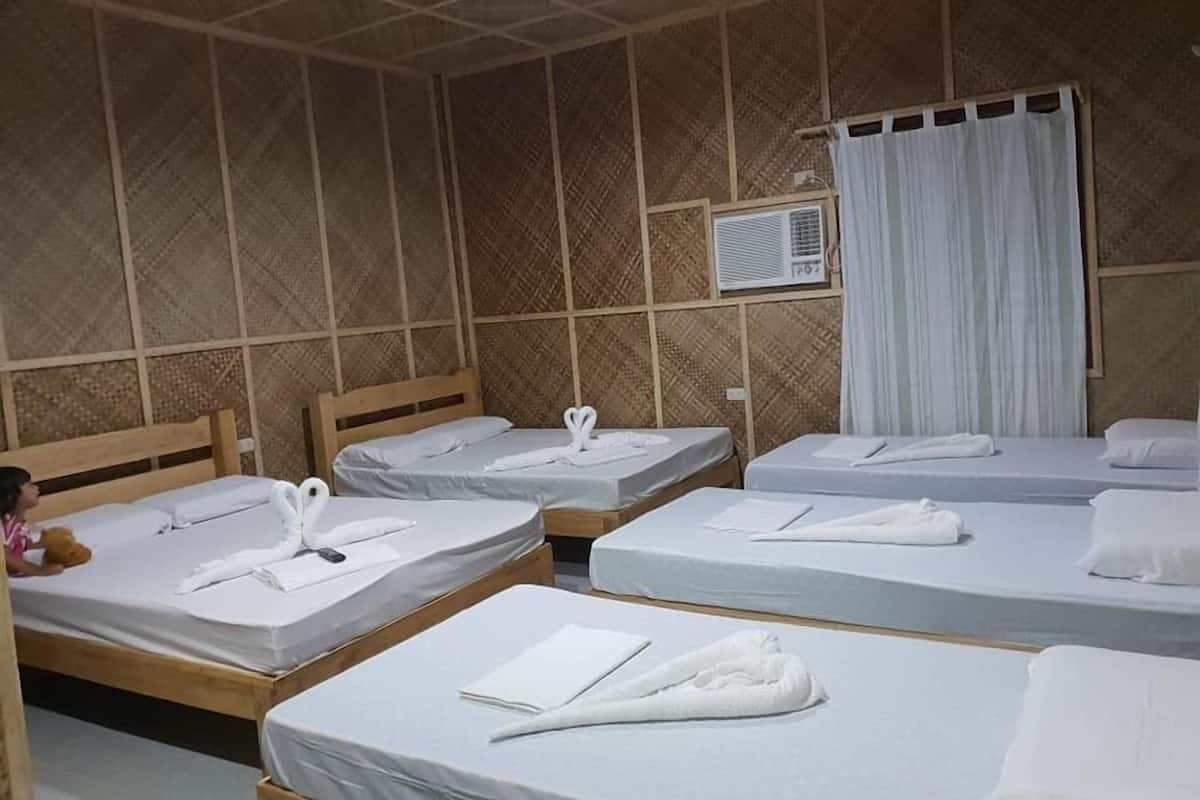 A room with four wooden beds, each with white linens and towels. There is a window with white curtains and an air conditioning unit on the wall.